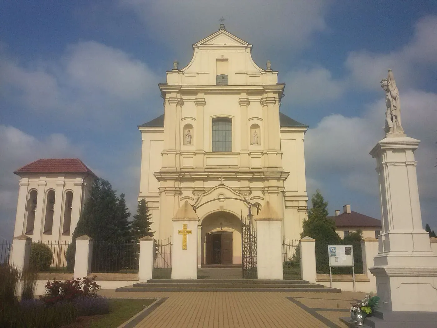 Jasnożółta, barokowa fasada z wolutowym szczytem i bramą wejściową, obok dzwonnica i kolumna z figurą, na tle pochmurnego nieba