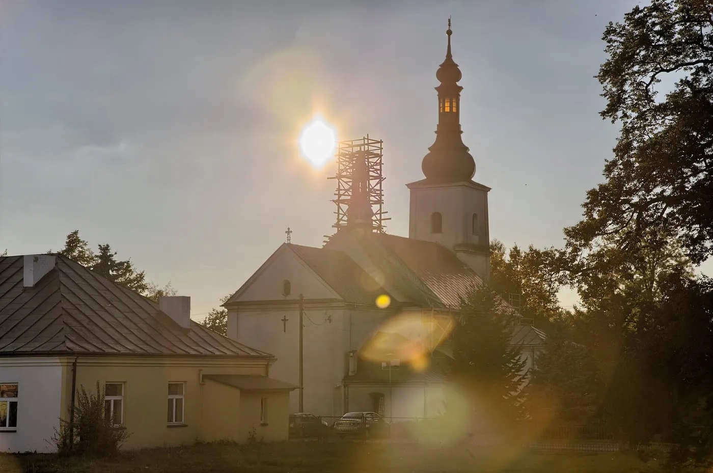 Kościół pod słońcem świecącym prosto w obiektyw: sylwetka wieży z rusztowaniem i cebulastym hełmem, obok parterowy budynek z blaszanym dachem