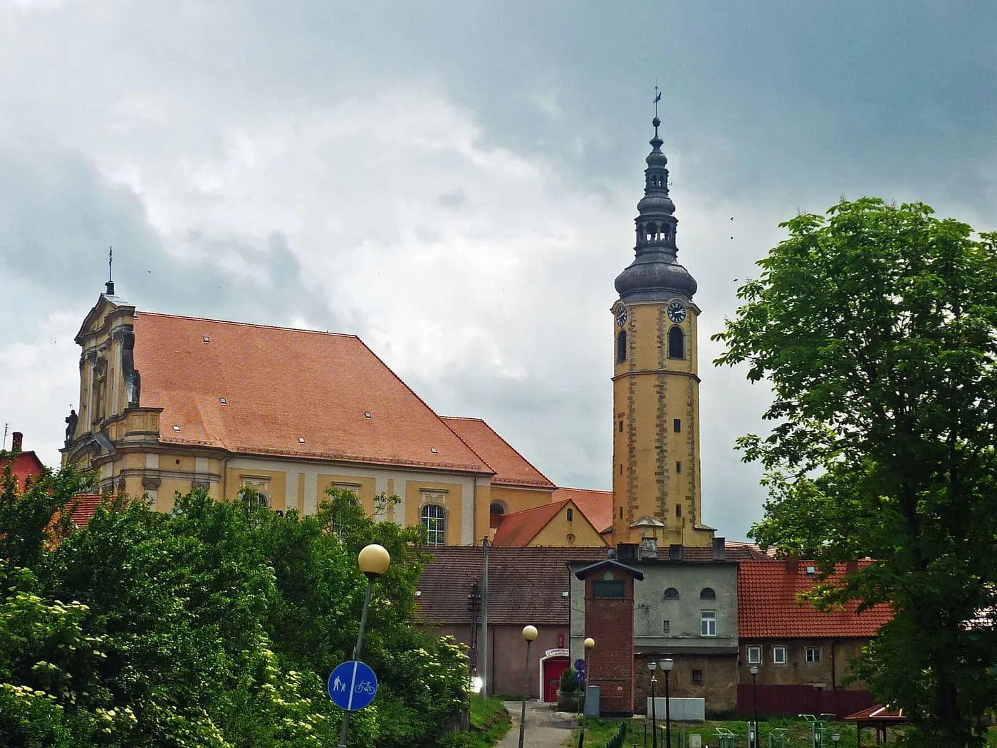 Barokowy kościół z pomarańczowym dachem i wysoką żółtą wieżą z zegarem, widziany zza drzew i zabudowań, pod pochmurnym niebem
