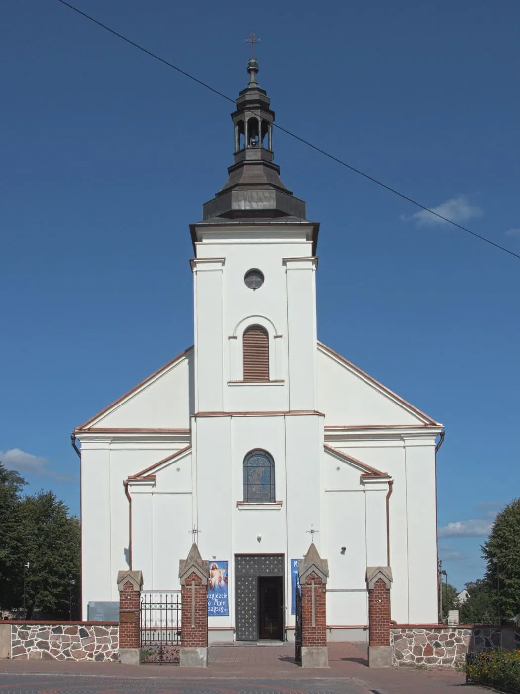 Biały kościół z jedną smukłą wieżą fasadową zwieńczoną ciemnym hełmem z latarnią, z ceglanymi słupkami bramy przed wejściem, na tle ciemnobłękitnego nieba