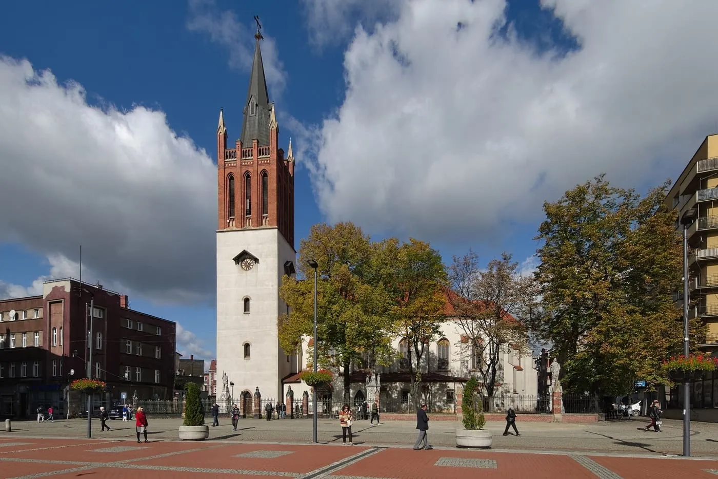 Kościół Wniebowzięcia NMP w Bytomiu z wieżą o białej, otynkowanej podstawie z zegarem i ceglanej, neogotyckiej górnej kondygnacji ze spiczastym hełmem, za jesiennymi drzewami, od strony placu