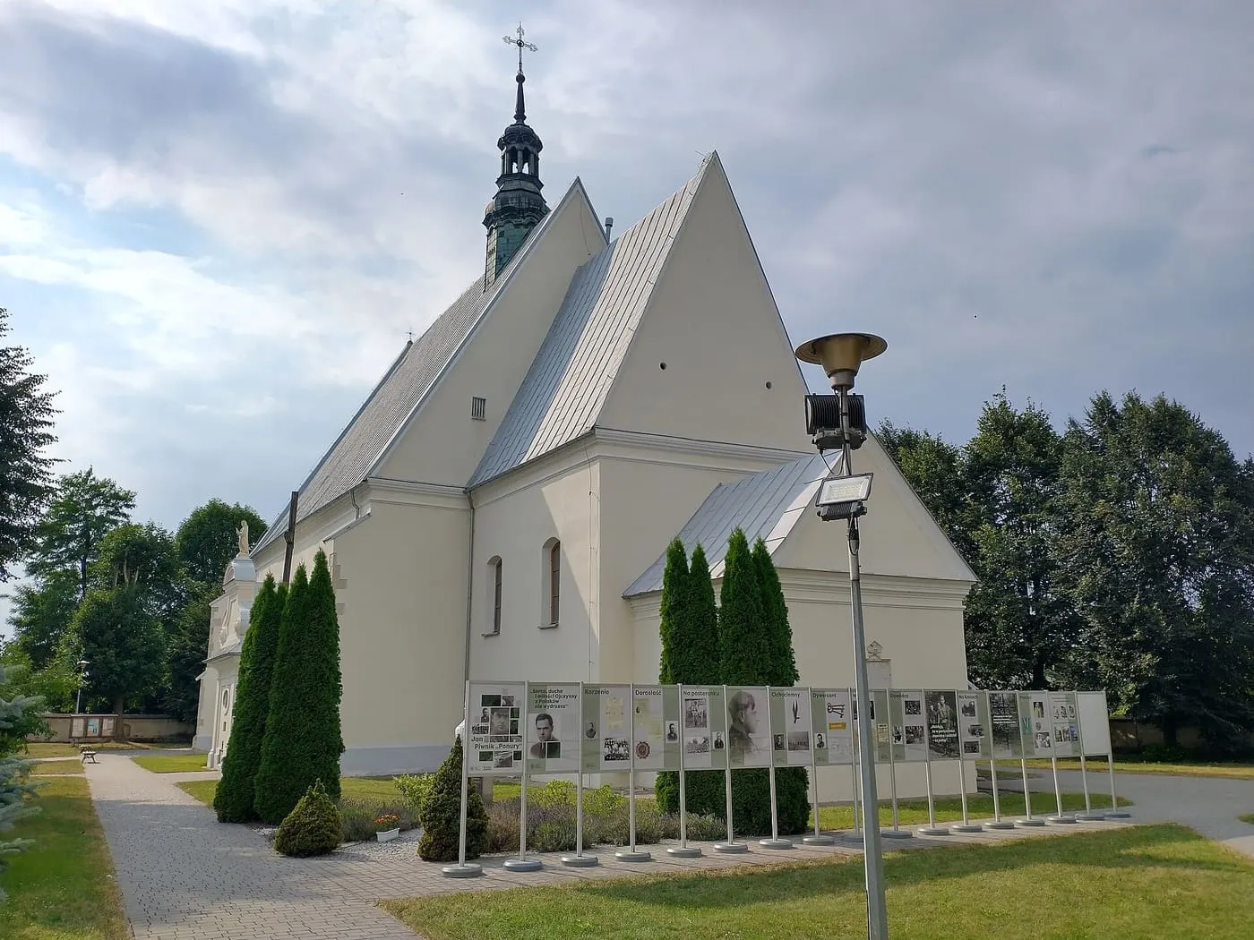 Biały, gotycki kościół z ostrymi szczytami i sygnaturką z zielonym hełmem, widziany od strony prezbiterium; przed nim rząd tablic wystawowych i strzyżone tuje