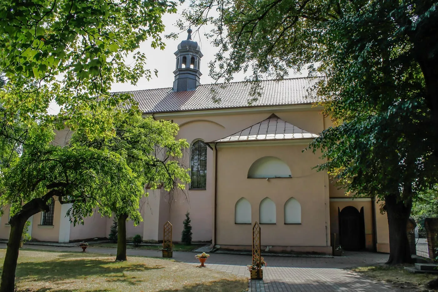 Różowo-kremowy kościół widziany z boku, z cebulastą wieżyczką, wśród drzew