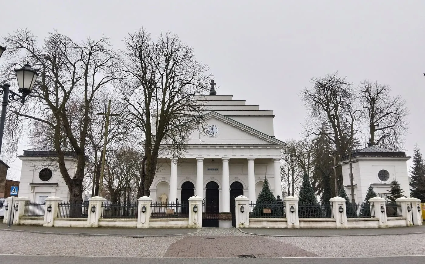 Klasycystyczny kościół Wniebowzięcia NMP w Kocku z kolumnowym portykiem i schodkowym szczytem, między dwiema ośmiobocznymi kaplicami, za żeliwnym ogrodzeniem