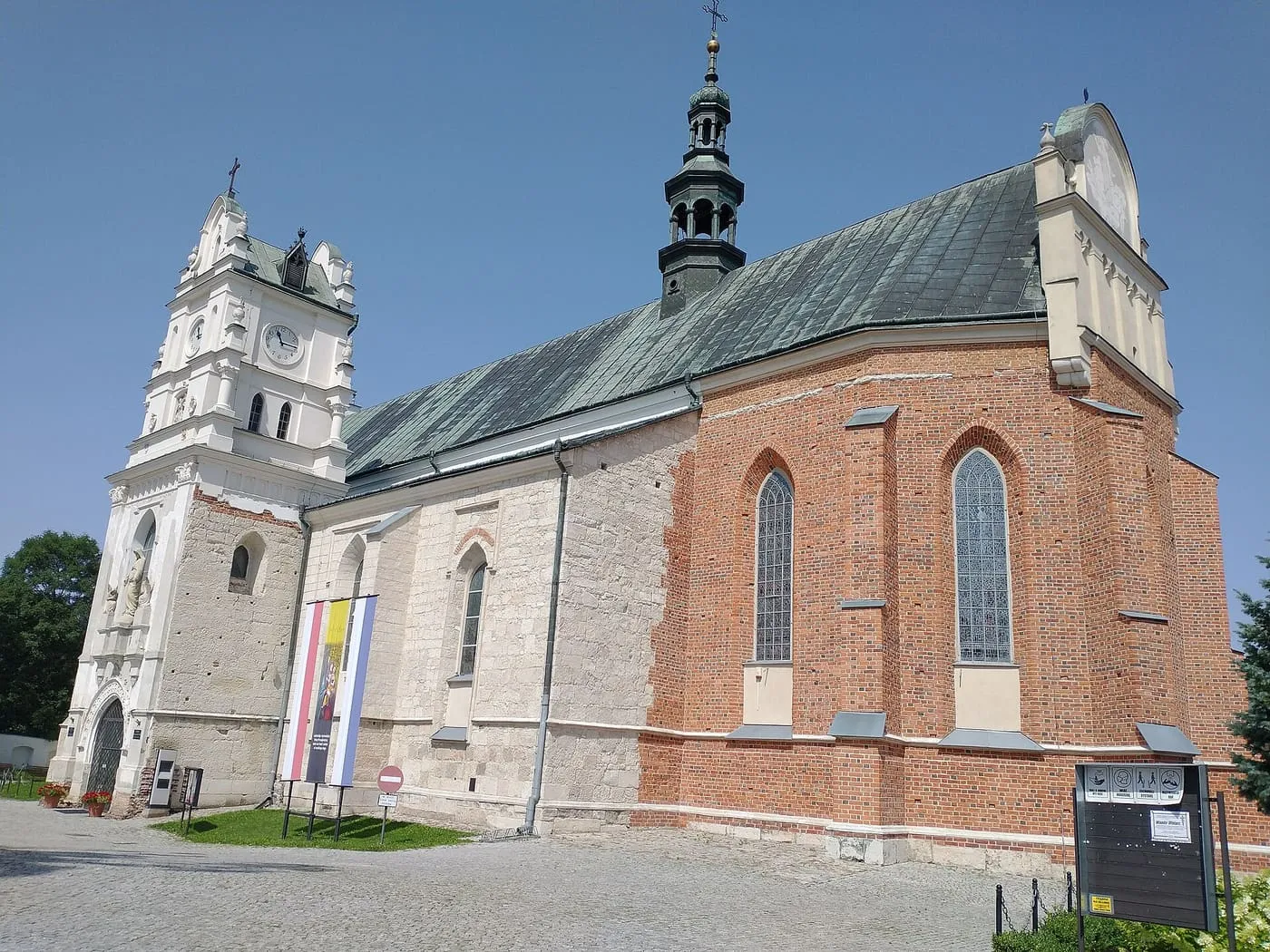 Kościół w Kraśniku z białą wieżą zegarową i ozdobną, zieloną wieżyczką nad ceglaną absydą, na tle błękitnego nieba