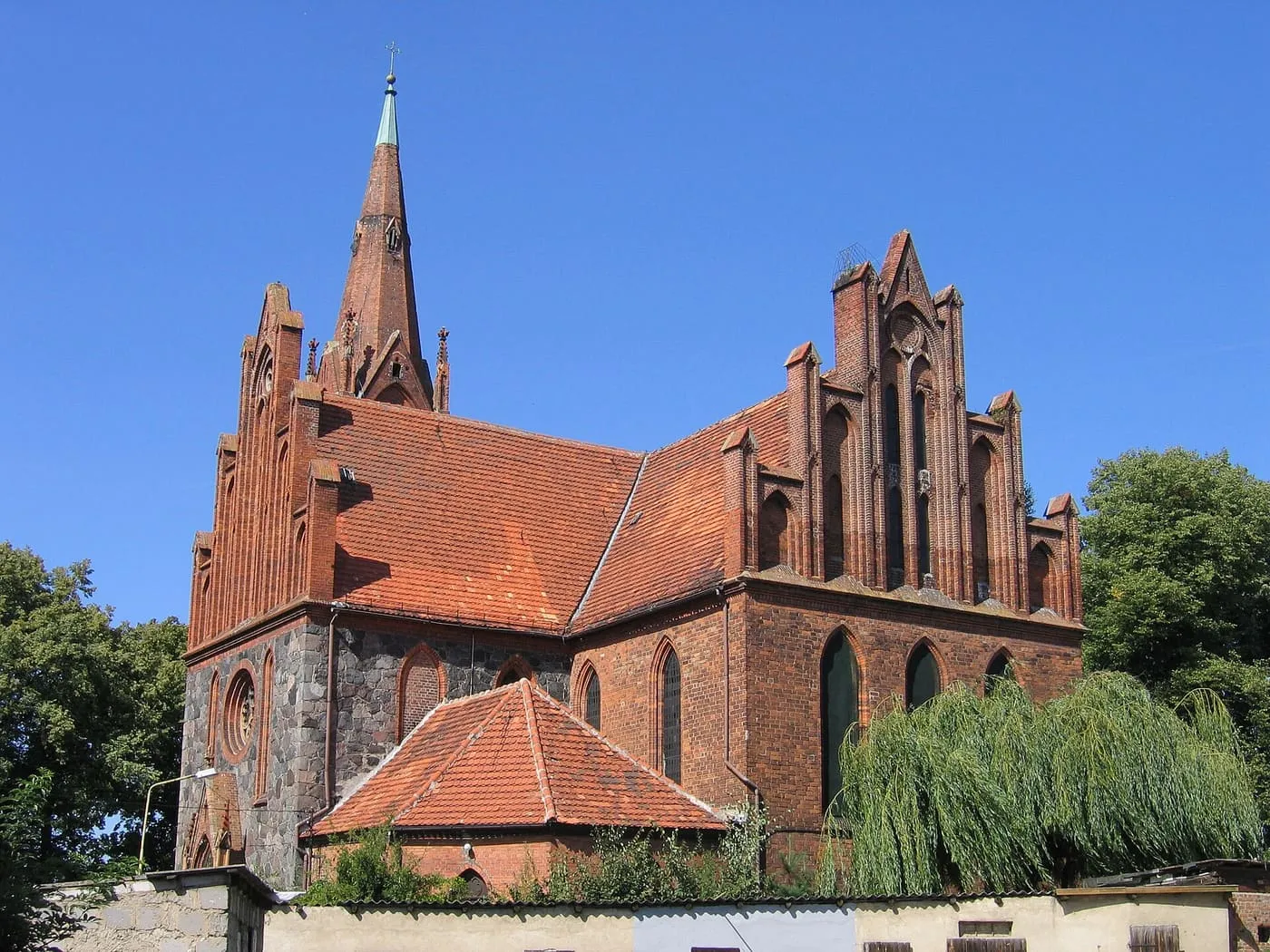 Ceglany, gotycki kościół z wysokimi, schodkowymi szczytami i strzelistą wieżyczką, z dolną częścią z polnych kamieni i czerwonym dachem, pod błękitnym niebem