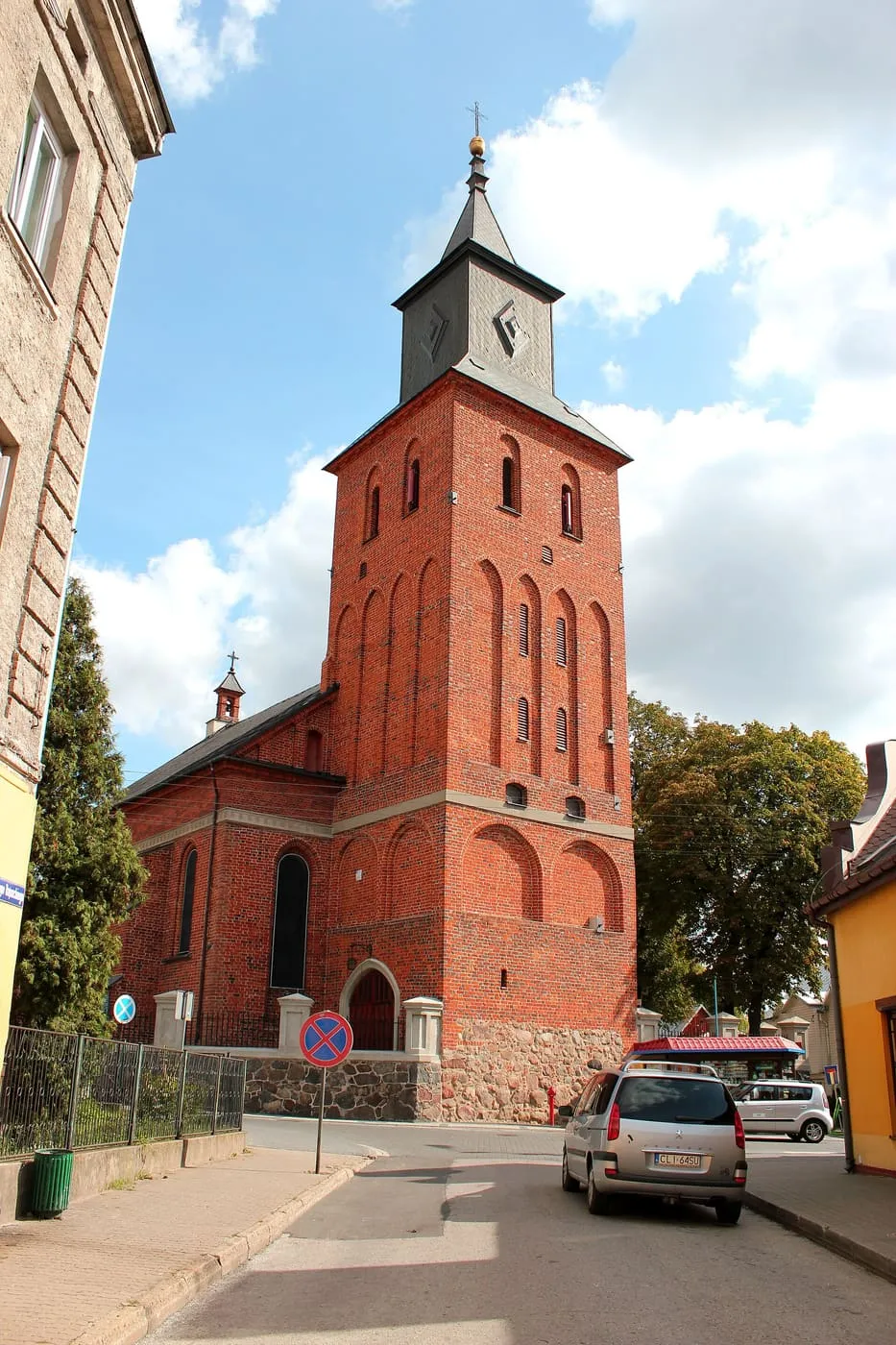 Ceglana wieża kościoła z wysokimi blendami i ciemnym, ostrosłupowym hełmem, obok niższa nawa; na pierwszym planie ulica z zaparkowanym samochodem