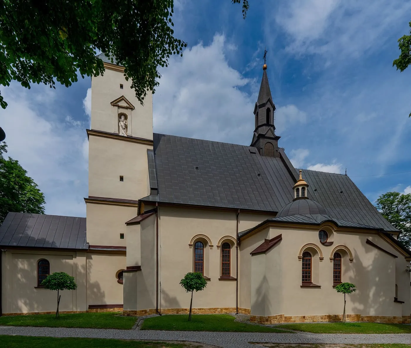 Kremowo-żółty, renesansowy kościół z ciemnym dachem, białą wieżą z figurą w niszy i smukłą sygnaturką, widziany zza gałęzi drzew, na tle błękitnego nieba
