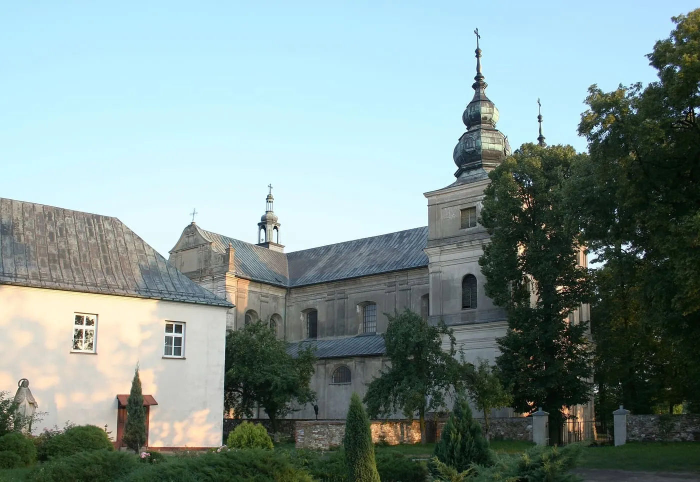 Szarobiały, barokowy kościół z wieżą zwieńczoną cebulastym hełmem, widziany zza zieleni i białego budynku o szarym dachu, w słoneczny dzień