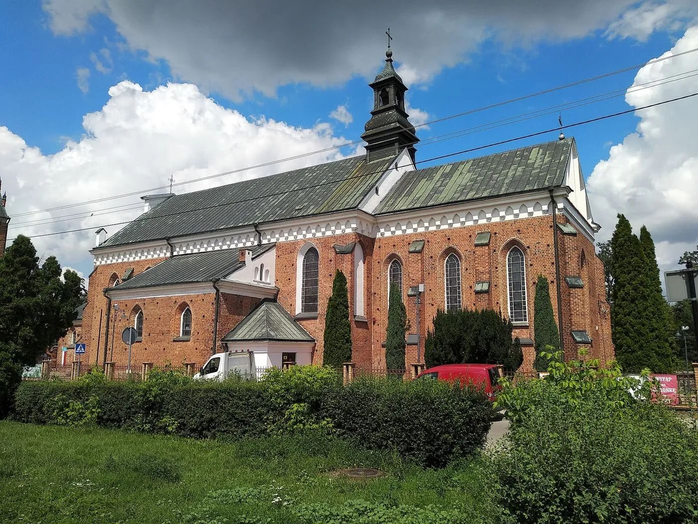 Ceglana, gotycka fara w Przasnyszu z białym fryzem z ostrołukowych blend pod okapem, zielonkawymi dachami i ciemną sygnaturką z latarnią, widziana zza żywopłotu