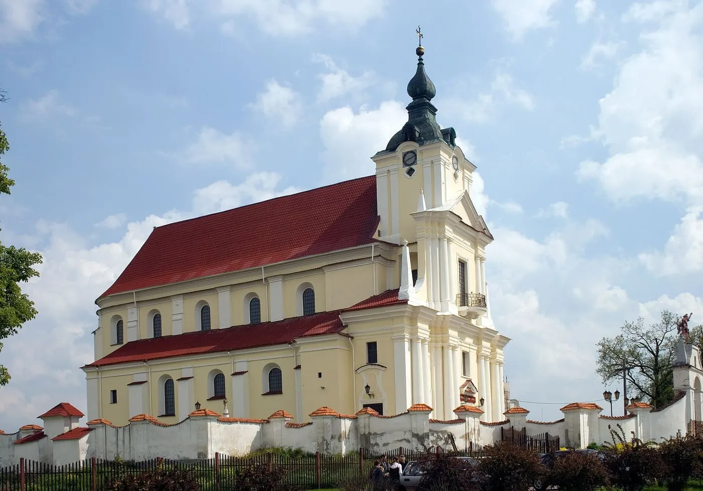 Barokowy kościół Wniebowzięcia NMP w Siemiatyczach o kremowych elewacjach, z czerwonym dachem i wieżą z zegarem zwieńczoną zielonym, cebulastym hełmem, za białym murem ogrodzenia
