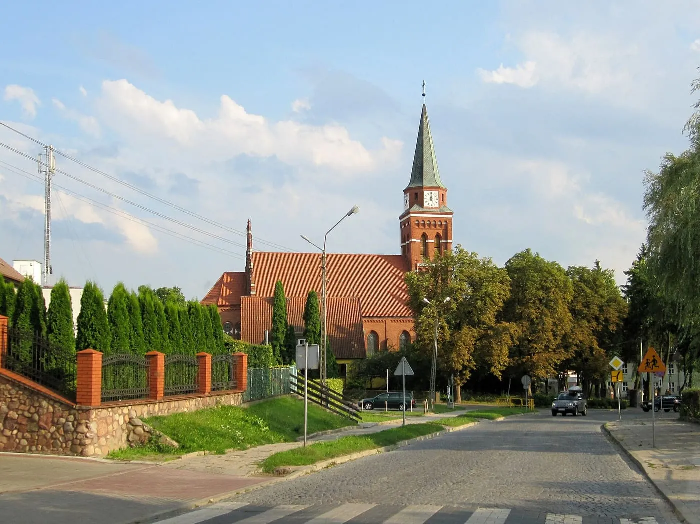 Ceglany, neogotycki kościół w Szczytnie z wysoką wieżą zwieńczoną smukłą, zieloną iglicą i zegarem, widziany znad ulicy obsadzonej żywopłotem z tui