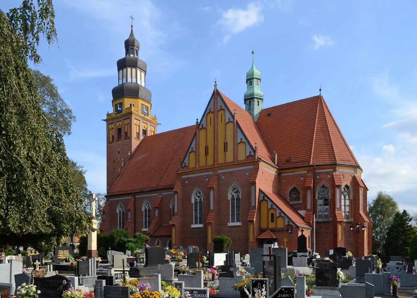 Neogotycki kościół w Wodzisławiu Śląskim z czerwonej i żółtej cegły, ze stromym szczytem i wieżą z ciemnym, bulwiastym hełmem, fotografowany znad cmentarza w pierwszym planie