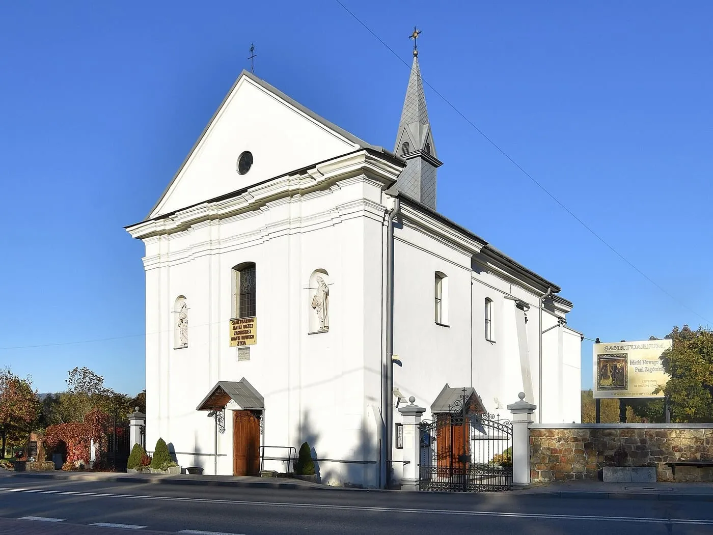 Biały, późnobarokowy kościół z trójkątnym szczytem fasady i smukłą wieżyczką z sygnaturką, za ogrodzeniem z bramką, przy ulicy; z prawej tablica informacyjna
