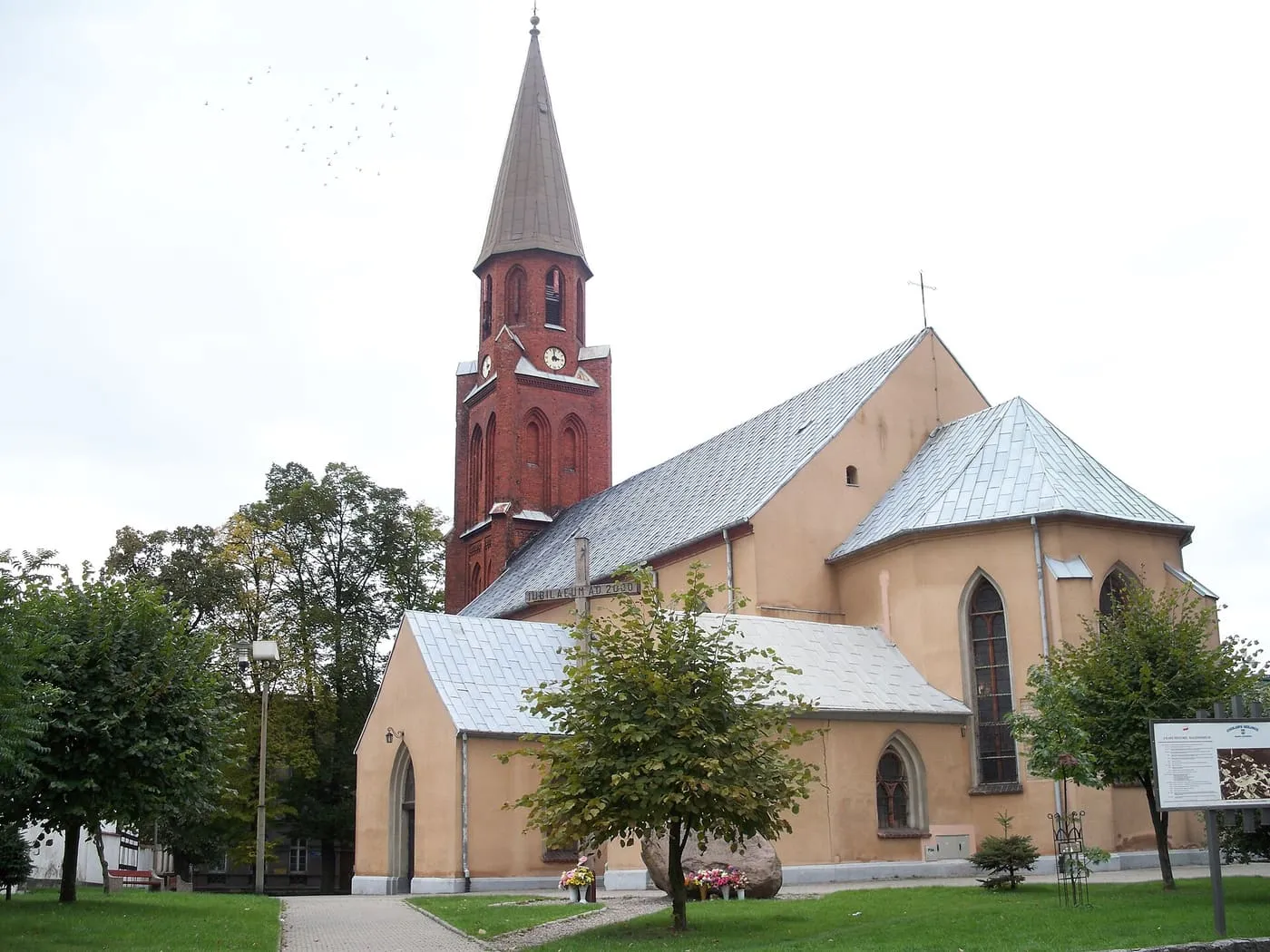 Kościół w kolorze piaskowym z ceglaną wieżą z zegarem i wysokim, szarym hełmem oraz jasnymi dachami; wokół trawnik i drzewa, na niebie stado ptaków