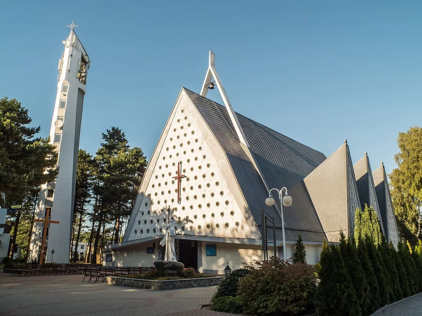 Nowoczesny, biały kościół o ostrych, trójkątnych dachach ozdobionych okrągłymi otworami, z wysoką, iglicową dzwonnicą i krzyżem, na tle błękitnego nieba