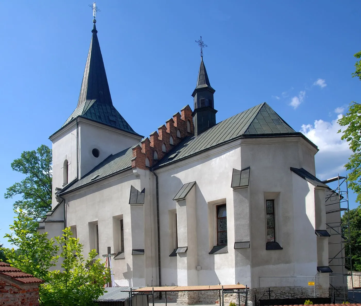 Biały kościół z bardzo ostrym, ciemnym hełmem wieży i ceglanym, krenelowanym wieńcem prezbiterium, na tle błękitnego nieba, po prawej stronie rusztowanie