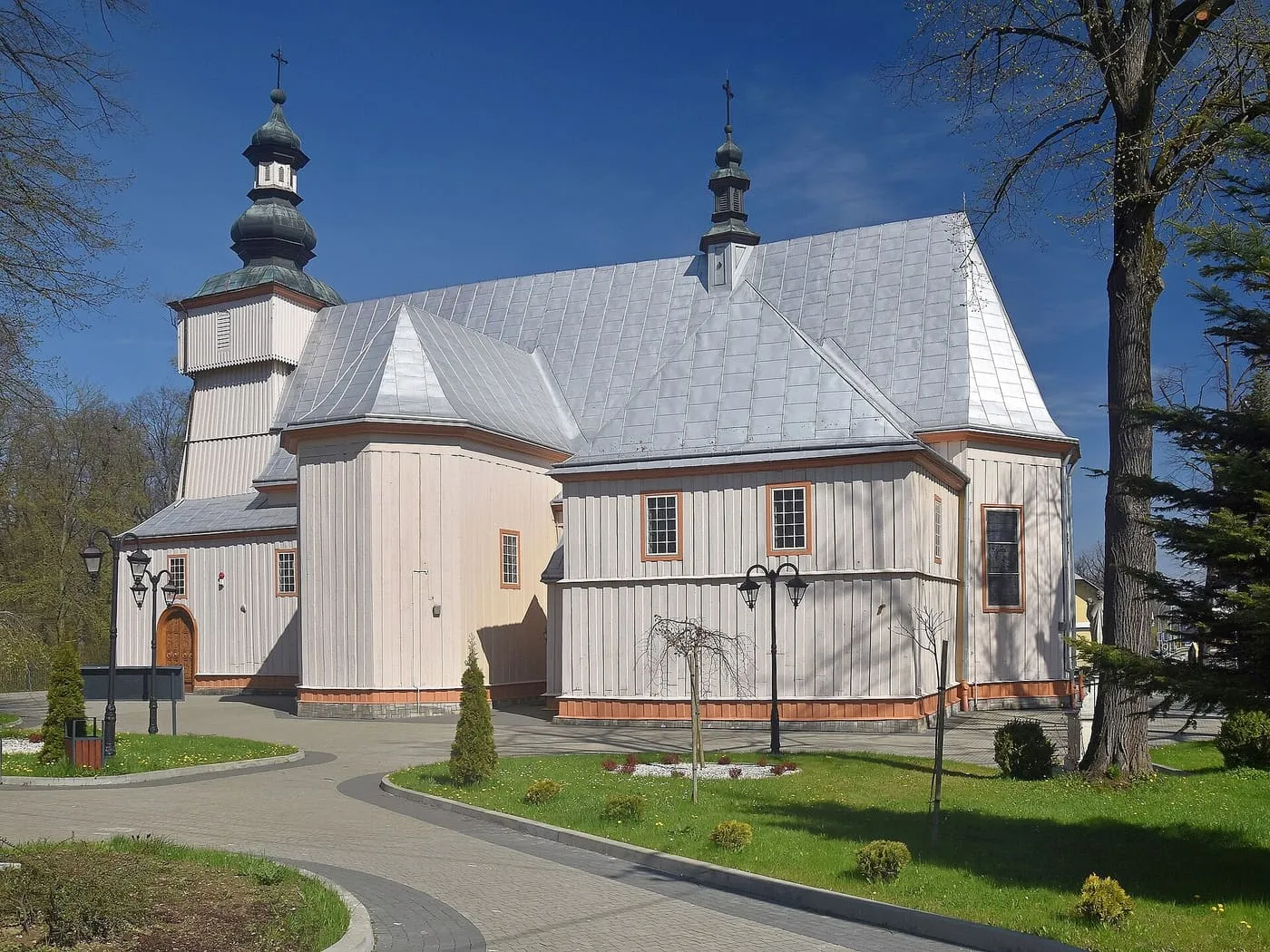 Drewniany kościół z jasnymi, pionowo szalowanymi ścianami i blaszanym dachem, z dwiema ciemnymi wieżyczkami o cebulastych hełmach; wokół trawniki, ścieżka i wysokie drzewa