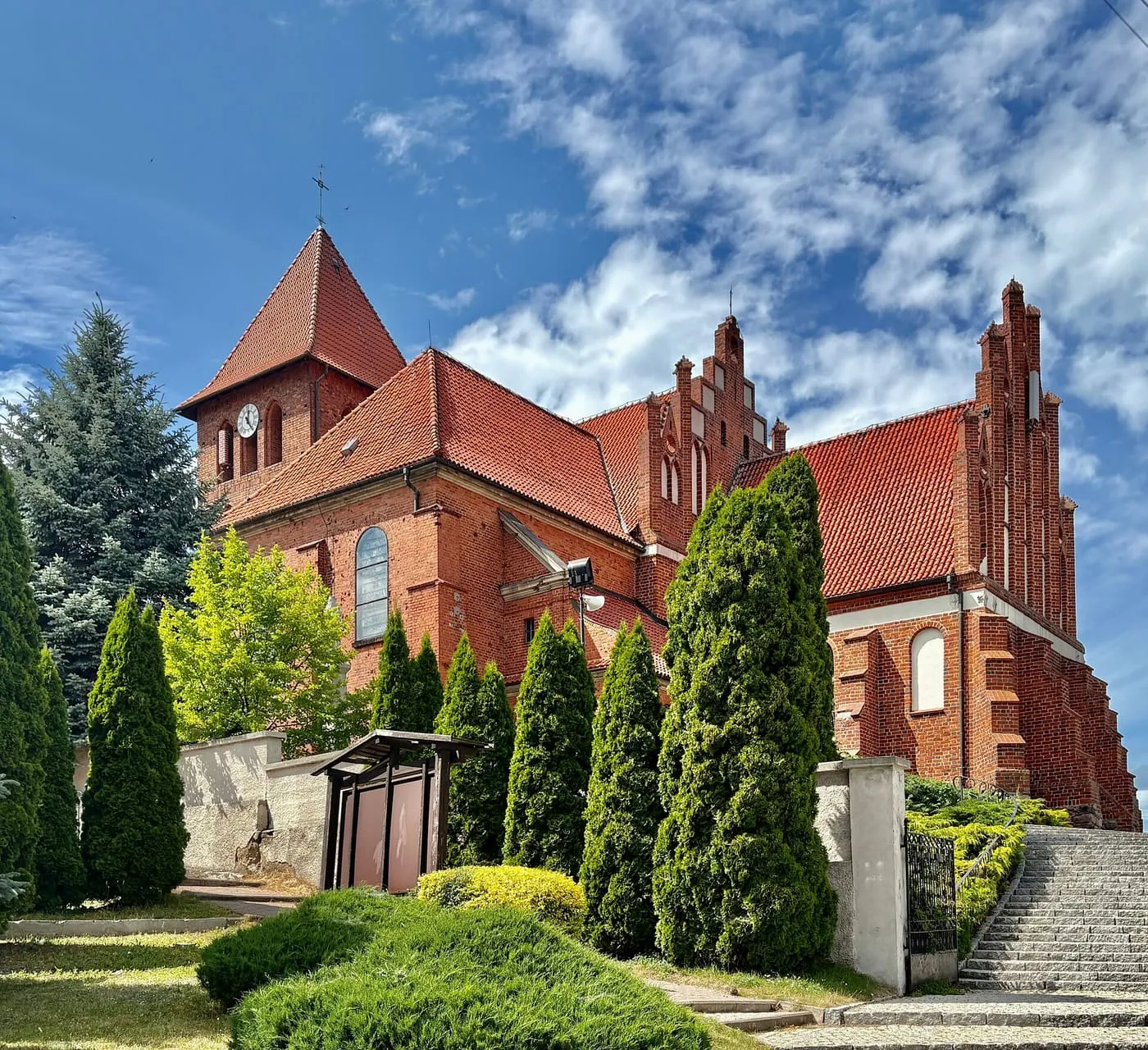 Ceglany kościół z czerwonymi dachami, schodkowymi szczytami i niską wieżą z zegarem, otoczony strzyżonymi tujami i zielenią, na tle błękitnego nieba z cienkimi chmurami