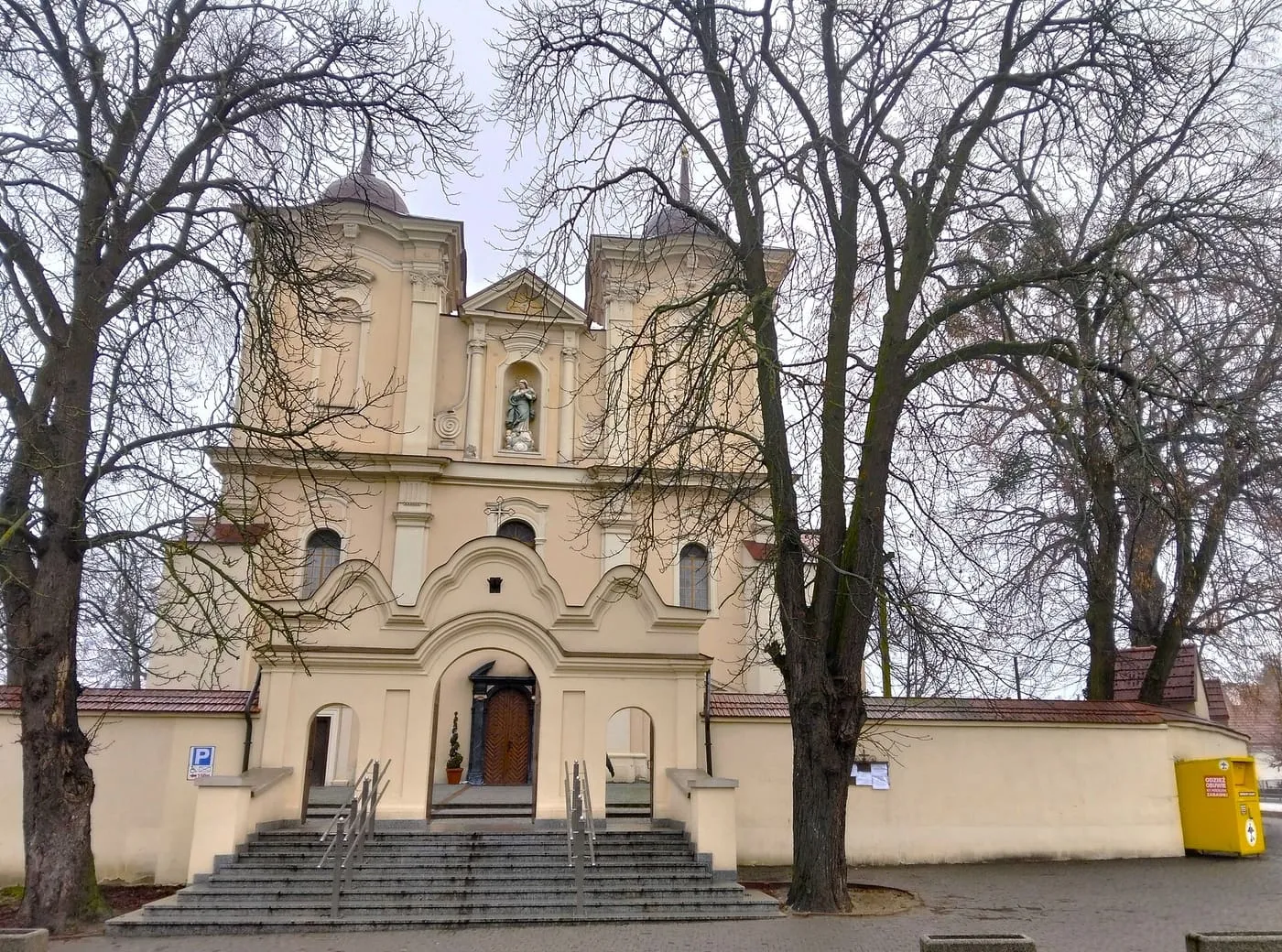 Jasnożółta, barokowa fasada z dwiema wieżami i schodami prowadzącymi do portalu, częściowo zasłonięta bezlistnymi drzewami, za murem otaczającym kościół