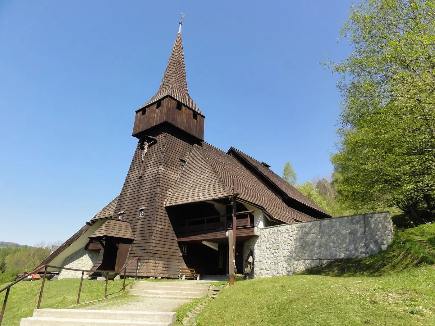Drewniany kościół pokryty ciemnym gontem, z wysoką, czworoboczną wieżą z ostrym hełmem, stojący na trawiastym stoku; do wejścia prowadzą betonowe schody