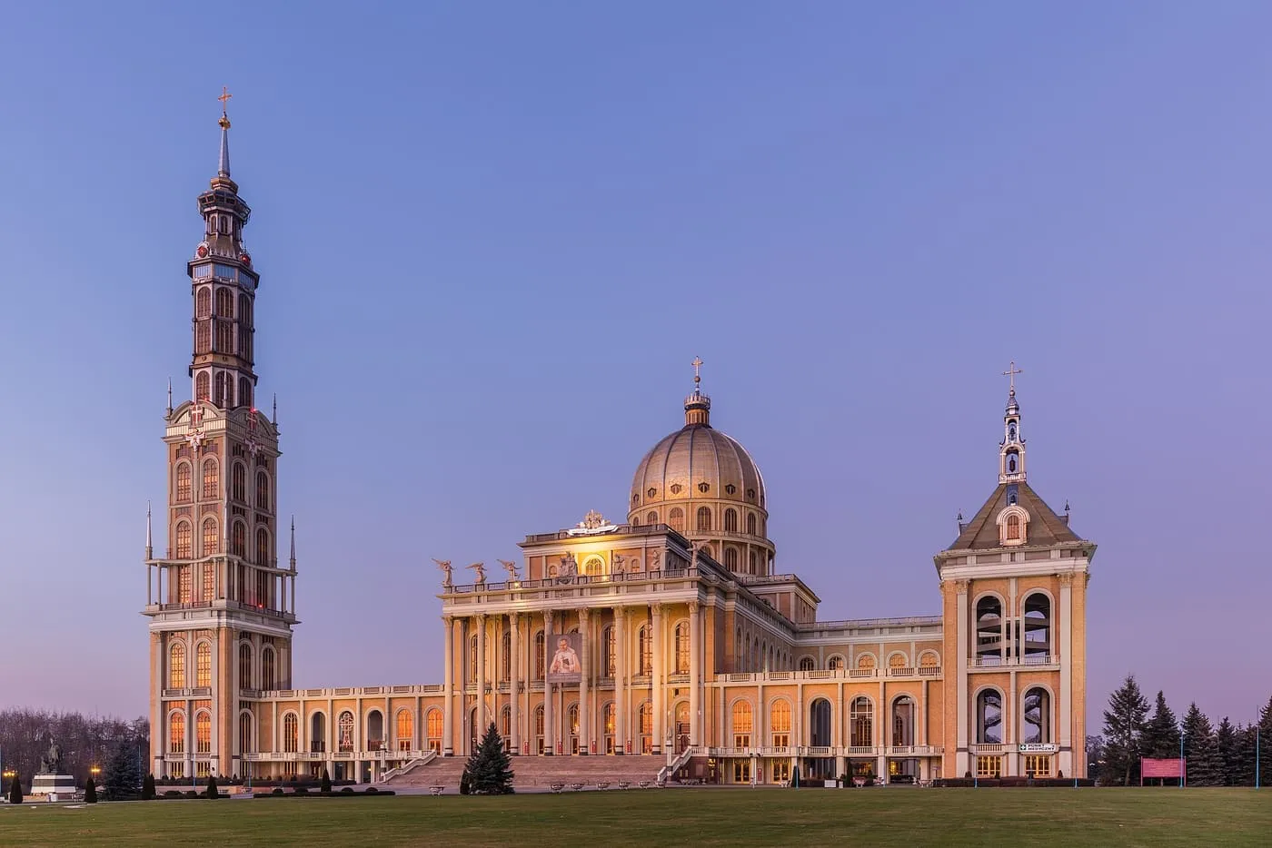 Bazylika licheńska o zmierzchu, rozświetlona złotym światłem: kopuła, kolumnowy portyk, wysoka wieża po lewej i dzwonnica po prawej, przed nią trawnik