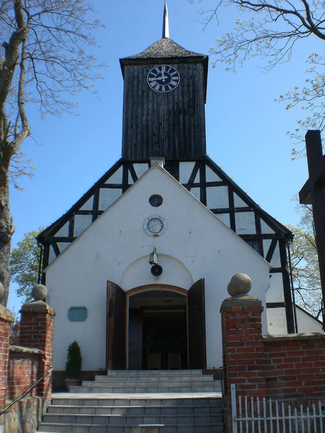 Biały szczyt kościoła z czarną, ryglową górną częścią i czarna drewniana wieża z zegarem, przed nią ceglane słupy bramy
