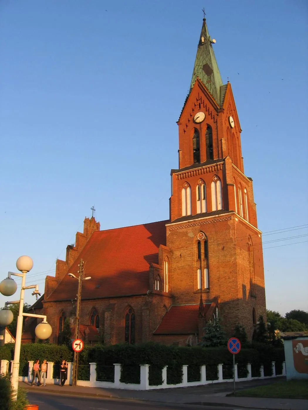 Ceglany, gotycki kościół z wysoką wieżą zwieńczoną zielonym hełmem, oświetlony ciepłym światłem zachodzącego słońca, przy ogrodzonej ulicy z lampami