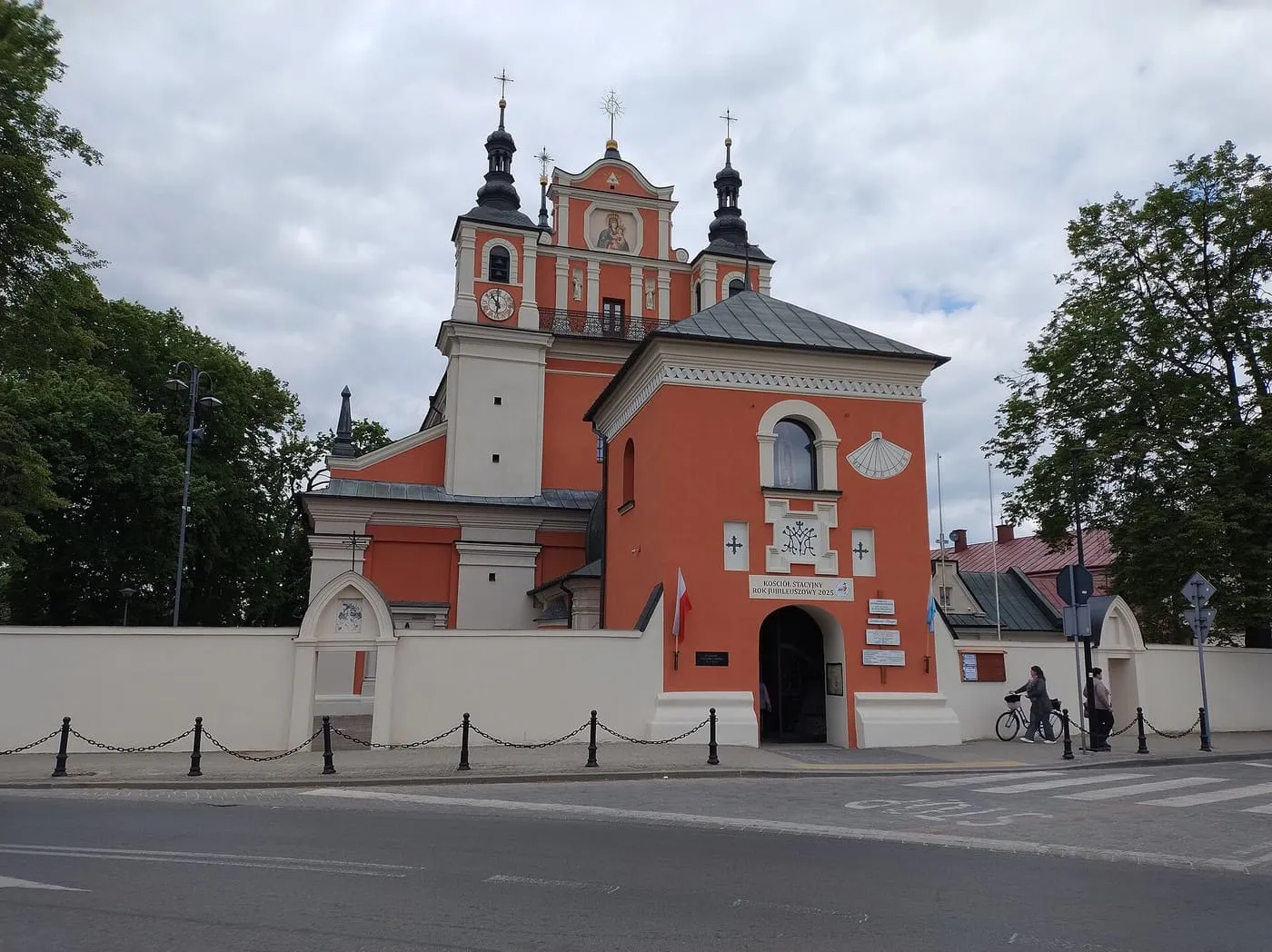 Pomarańczowa, barokowa fasada sanktuarium w Janowie Lubelskim z dwiema ciemnymi kopułami i wizerunkiem Matki Bożej