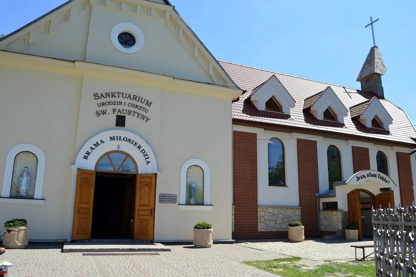 Kremowa fasada z napisami „Sanktuarium urodzin i chrztu św. Faustyny” i „Brama Miłosierdzia”, a obok ceglana, nowsza część kościoła z dachem z lukarnami