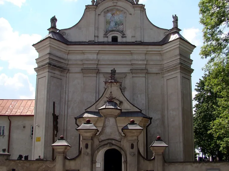 Szara, barokowa fasada sanktuarium w Krasnobrodzie z bramą wjazdową i napisem „Sanktuarium Maryjne”