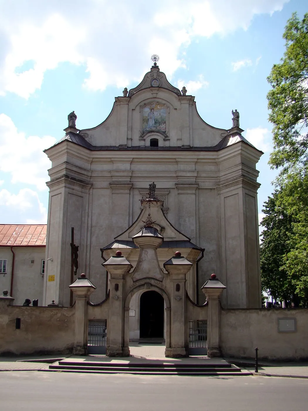 Szara, barokowa fasada sanktuarium w Krasnobrodzie z bramą wjazdową i napisem „Sanktuarium Maryjne”