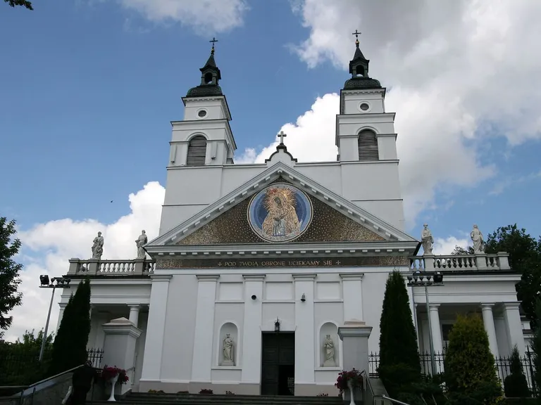 Biała, klasycystyczna fasada kolegiaty św. Antoniego z dwiema wieżami i mozaiką Matki Bożej w trójkątnym przyczółku, pod którą widnieje napis „Pod Twoją obronę uciekamy się”; przed wejściem schody i kwiaty