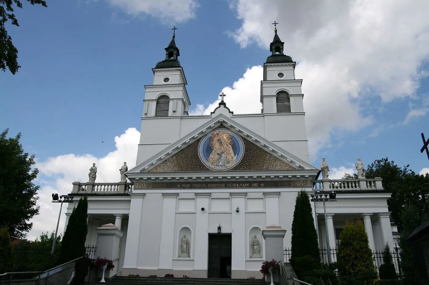 Biała, klasycystyczna fasada kolegiaty św. Antoniego z dwiema wieżami i mozaiką Matki Bożej w trójkątnym przyczółku, pod którą widnieje napis „Pod Twoją obronę uciekamy się”; przed wejściem schody i kwiaty