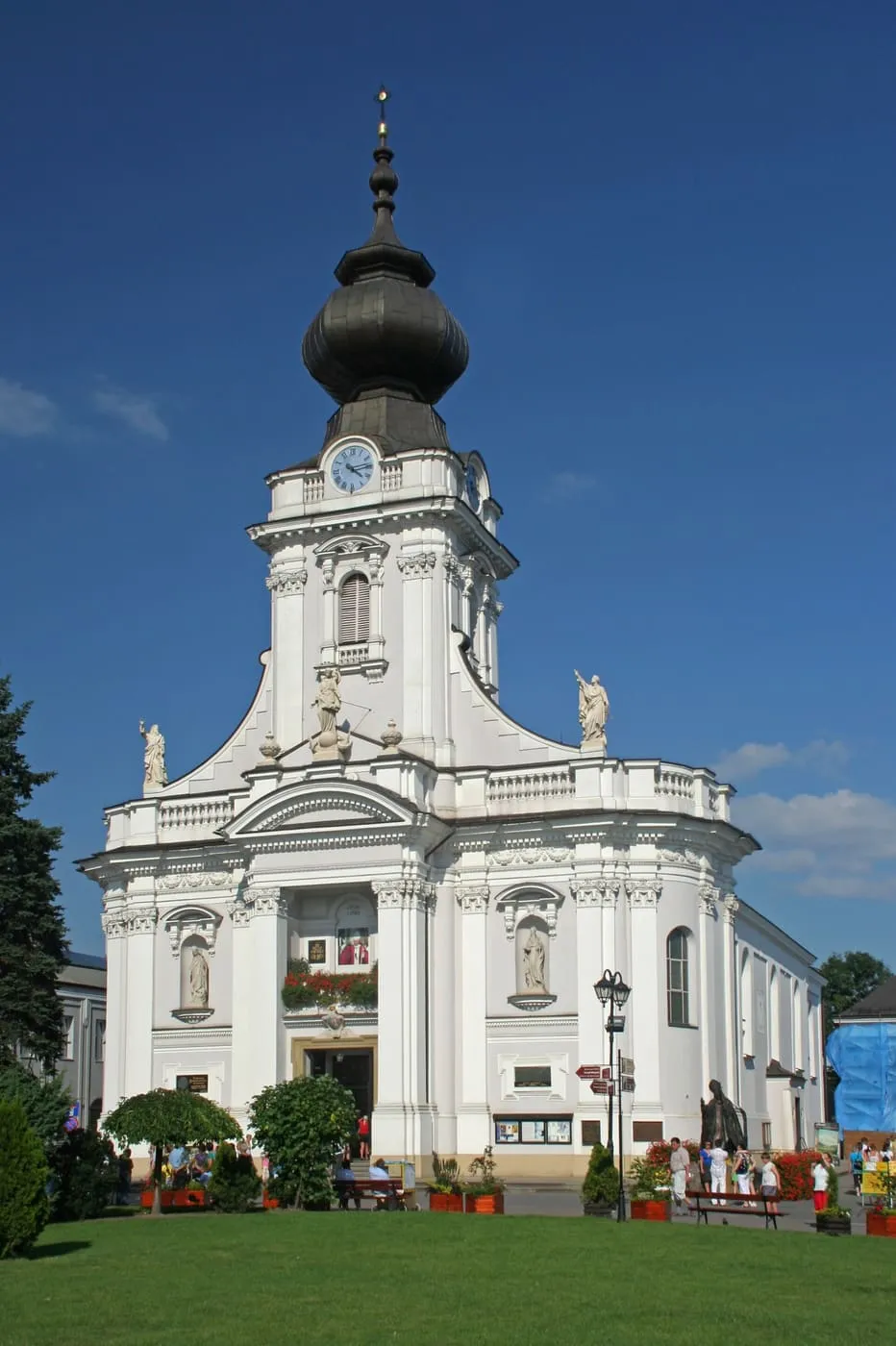 Biała, późnobarokowa fasada bazyliki w Wadowicach z wieżą zwieńczoną czarnym, baniastym hełmem i zegarem, figurami świętych we wnękach i portretem Jana Pawła II nad wejściem