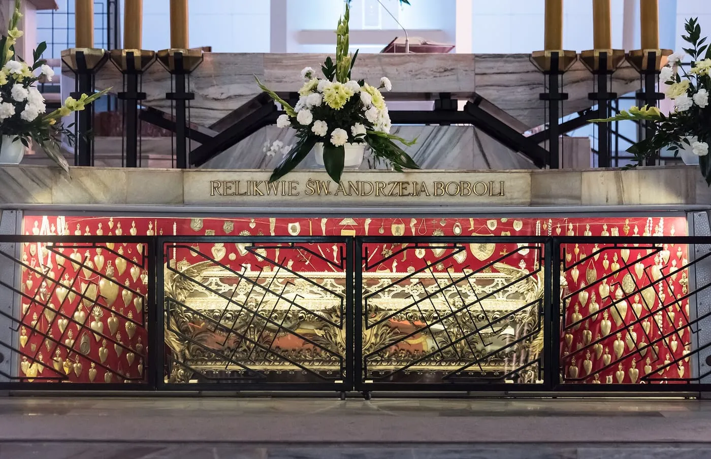 Relikwie św. Andrzeja Boboli w ołtarzu sanktuarium w Warszawie: pod napisem „Relikwie św. Andrzeja Boboli” w przeszklonej gablocie za kratą stoi złocony relikwiarz, a wokół niego na czerwonym tle wiszą złote wota w kształcie serc, na górze płoną świece i stoją bukiety białych kwiatów