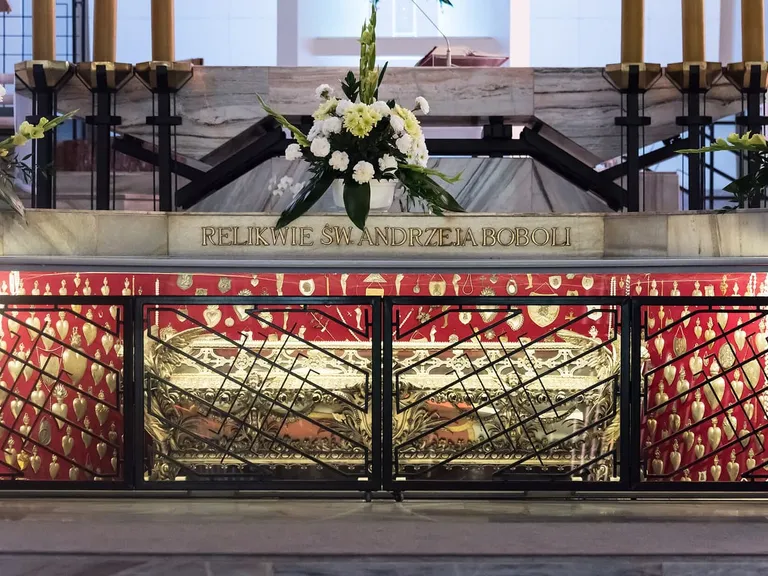Relikwie św. Andrzeja Boboli w ołtarzu sanktuarium w Warszawie: pod napisem „Relikwie św. Andrzeja Boboli” w przeszklonej gablocie za kratą stoi złocony relikwiarz, a wokół niego na czerwonym tle wiszą złote wota w kształcie serc, na górze płoną świece i stoją bukiety białych kwiatów