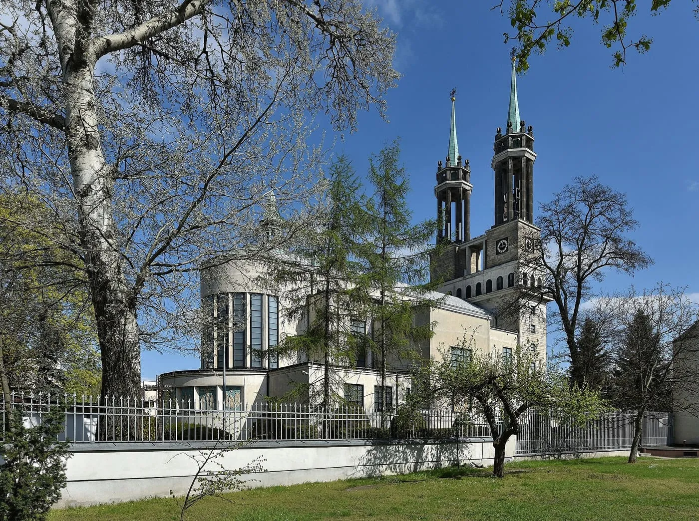Kościół św. Stanisława Kostki na warszawskim Żoliborzu widziany od strony prezbiterium: jasny, modernistyczny budynek z okrągłą apsydą i dwiema smukłymi wieżami z zielonymi hełmami, przed nim żelazne ogrodzenie i drzewa