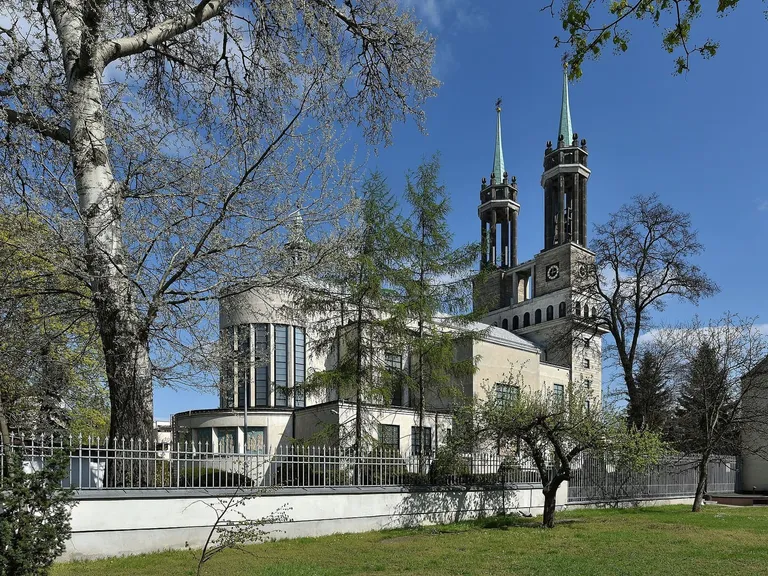 Kościół św. Stanisława Kostki na warszawskim Żoliborzu widziany od strony prezbiterium: jasny, modernistyczny budynek z okrągłą apsydą i dwiema smukłymi wieżami z zielonymi hełmami, przed nim żelazne ogrodzenie i drzewa